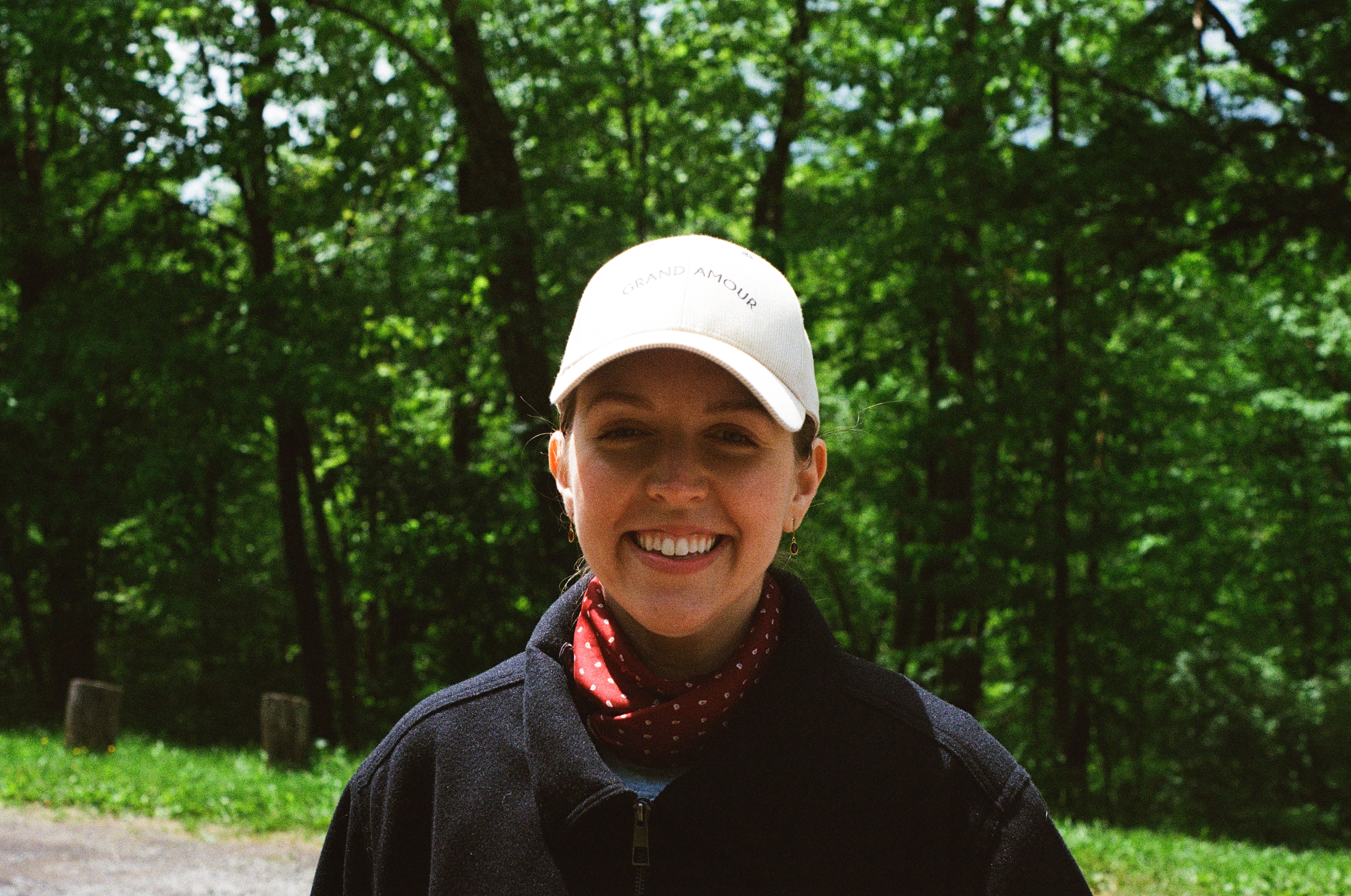 The photograph depicts a woman standing in front of a backdrop of lush trees and inviting grass. She is attired in a white...